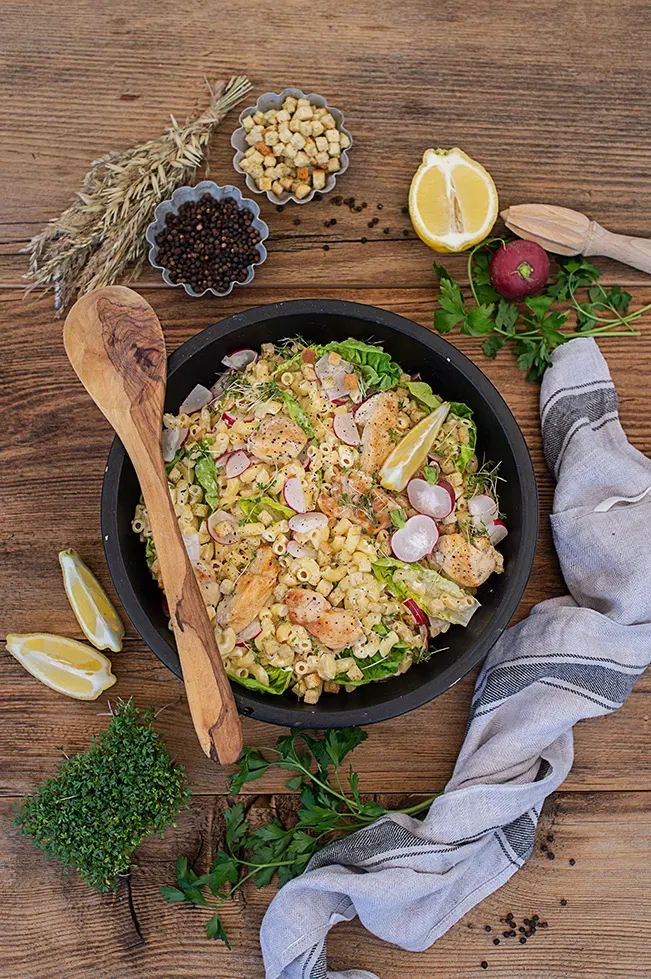Caesar Salad mit Nudeln, Huhn, Radieschen und Zitronenscheiben in schwarzer Schüssel auf Holztisch
