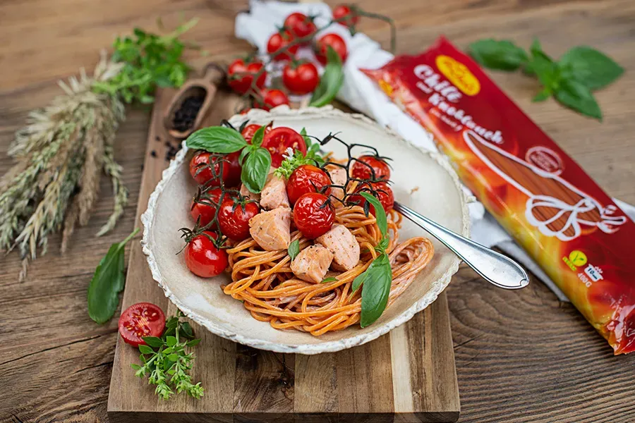 Chili-Knoblauch-Nudelbowl mit Lachs, Spaghetti, Kirschtomaten und Kräutern auf einem Holzbrett neben der Verpackung