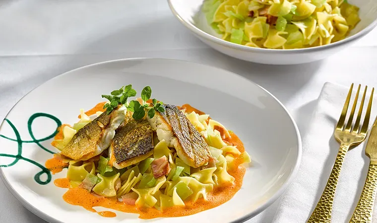 Donauzander mit Speckkrautfleckerl und Paprikaschaum, Pasta mit gebratenem Fisch auf weißem Tisch mit Besteck