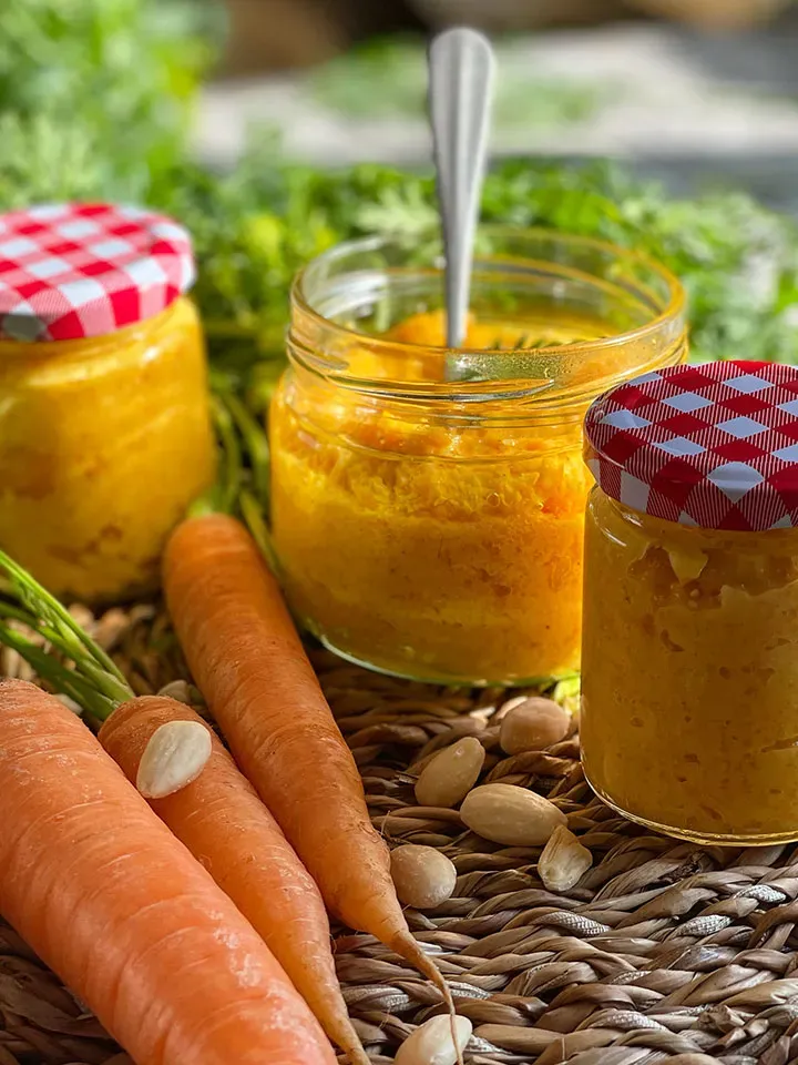 Karottenpesto in Gläsern neben frischen Karotten und Mandeln auf einem Tisch