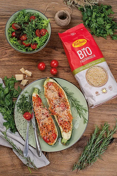 Gefüllte Zucchinischiffchen mit Tomaten, Käse, Salat und Bio-Fadennudeln auf Holztisch