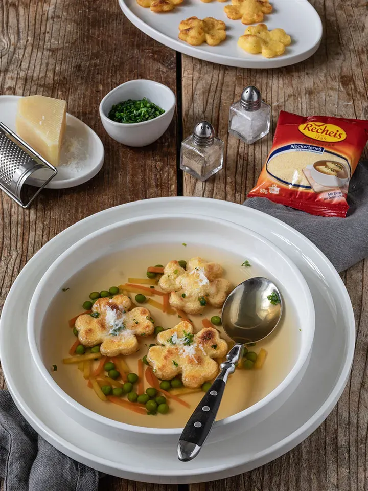 Glückssuppe mit Grießschöberl in Blumenform und Gemüse in einer weißen Schüssel auf Holztisch