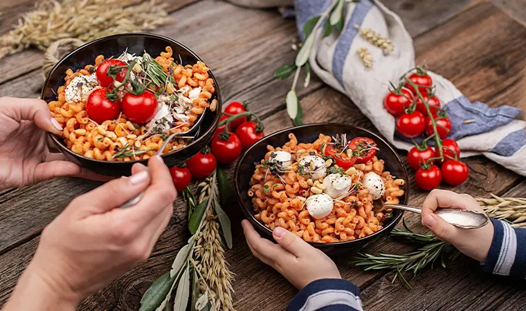Zwei Hände halten Schalen mit Goldlöckchen Minis in Tomaten-Ricotta-Sauce, dazu frische Tomaten, auf Holztisch