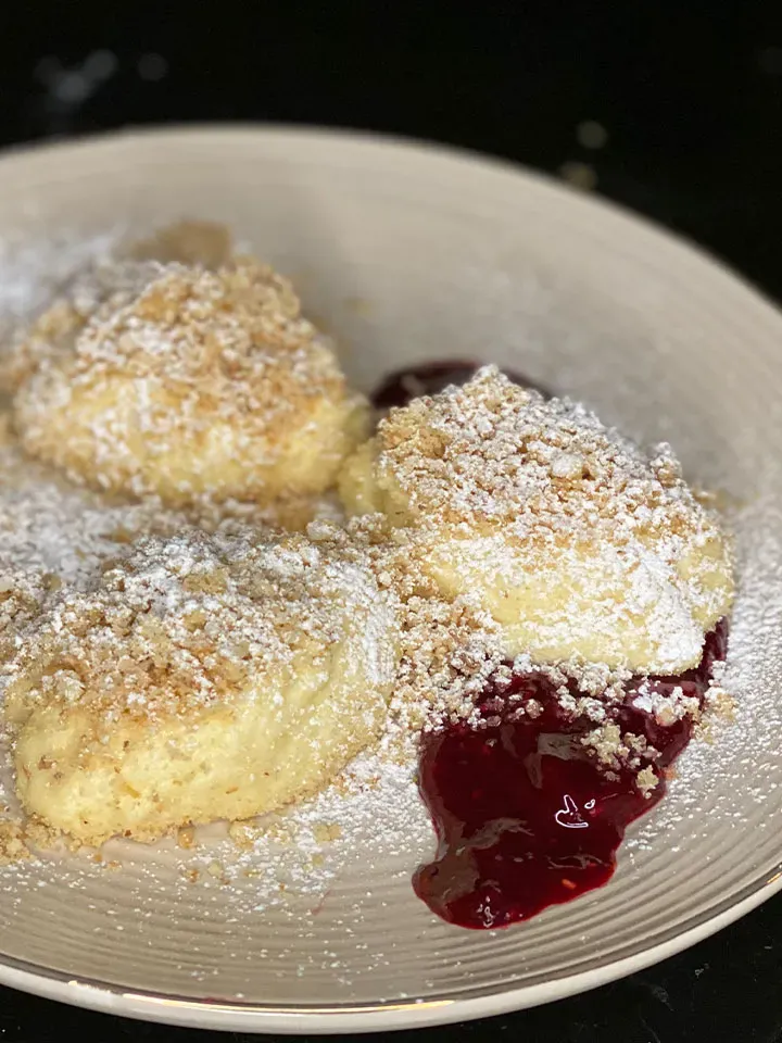 Drei süße Grieß-Topfen-Nockerl mit Nussbröseln, Puderzucker und roter Fruchtsauce auf einem Teller
