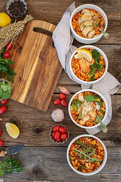 Hörnchen mit Tomatensauce und Huhn als Meal Prep: vier Schalen mit Pasta und Zutaten auf Holztisch mit Schneidebrett