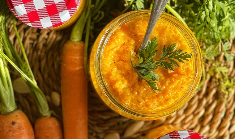 Karottenpesto in einem Glas neben frischen Karotten auf einem geflochtenen Untergrund