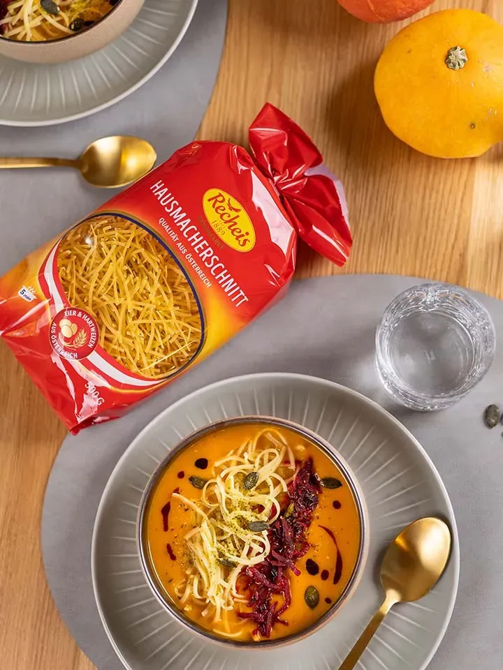 Kürbiscremesuppe mit Hausmacherschnitt-Fadennudeln und Kürbiskernen auf grauem Teller mit goldfarbenem Löffel