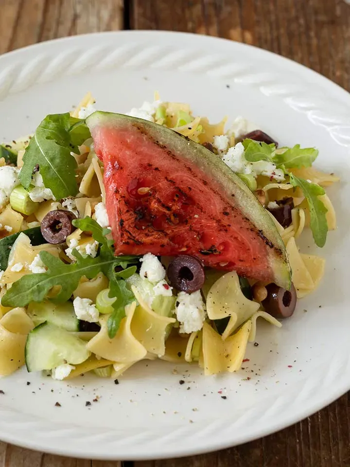 Salat mit gegrillter Wassermelone, Nudeln, Oliven, Rucola und Feta auf weißem Teller