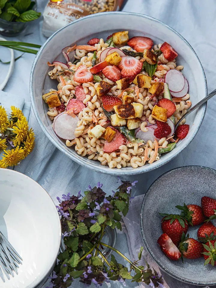 Muttertagspicknick: Schüssel Nudelsalat mit Spargel, Erdbeeren, Radieschen und gebratenem Käse, daneben Blumen