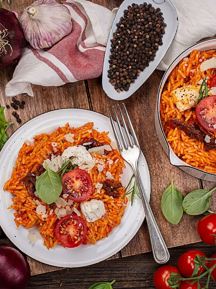 Nudel-Risotto mit Tomaten, Mozzarella und Basilikum auf Teller auf Holztisch mit Gewürzen