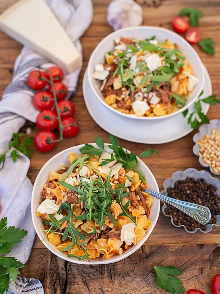Nudelbowl mit Pulled Beef: zwei Schalen Pasta mit Rucola, Käse und Tomaten auf Holztisch