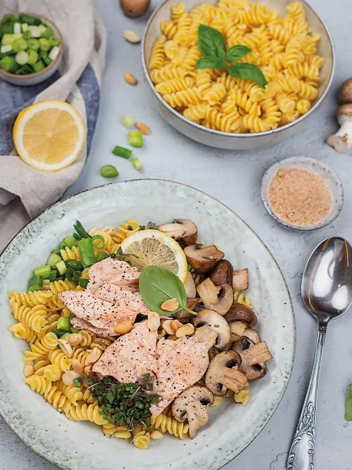 Nudeln mit Lachs und Champignons, Frühlingszwiebeln und Zitronenscheibe auf Teller auf Tisch