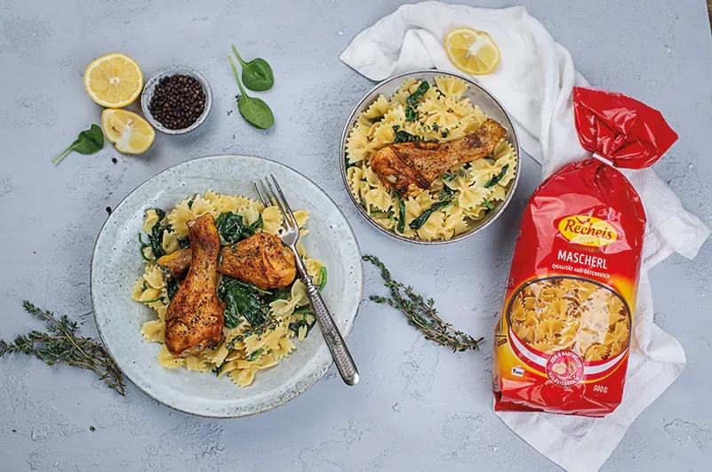 Nudelpfanne mit Hähnchen und Spinat: zwei Teller Farfalle mit Hähnchenschenkeln neben Nudelpackung und Zitronen