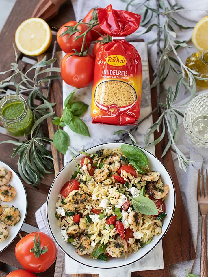 Schüssel mit Nudelreis, Garnelen, Tomaten und Kräutern neben Nudelreis und frischen Zutaten