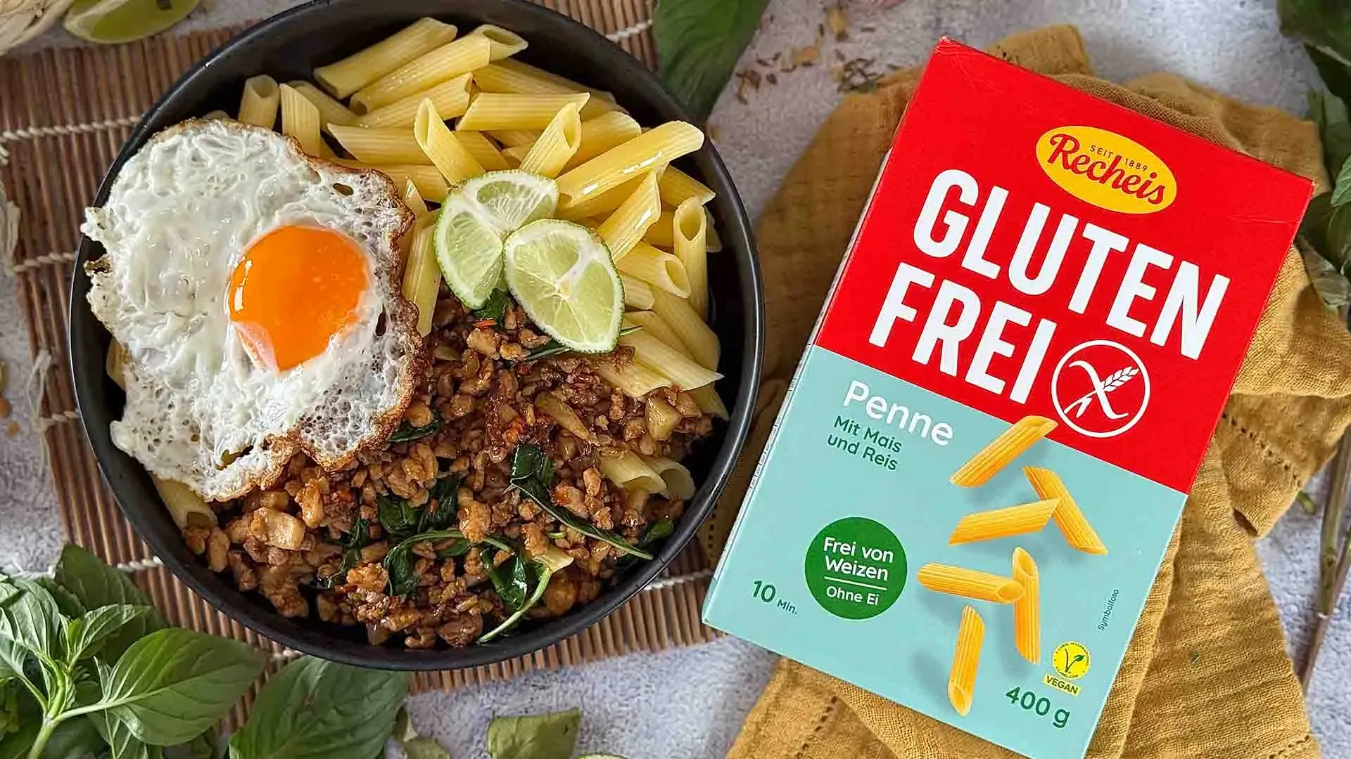 Pad Kra Pao mit Penne, Spiegelei, Hackfleisch und Limettenscheiben in einer Schüssel neben der Packung glutenfreier Penne