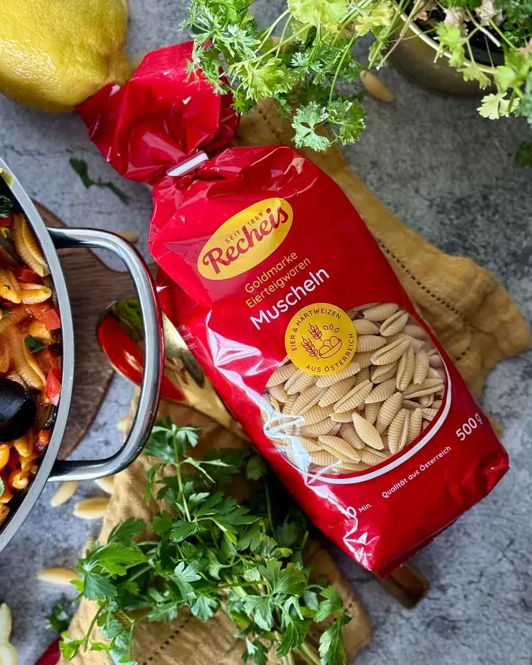 Paella mit Nudeln: rote Packung Muschelnudeln neben frischen Kräutern und einem Topf mit Essen