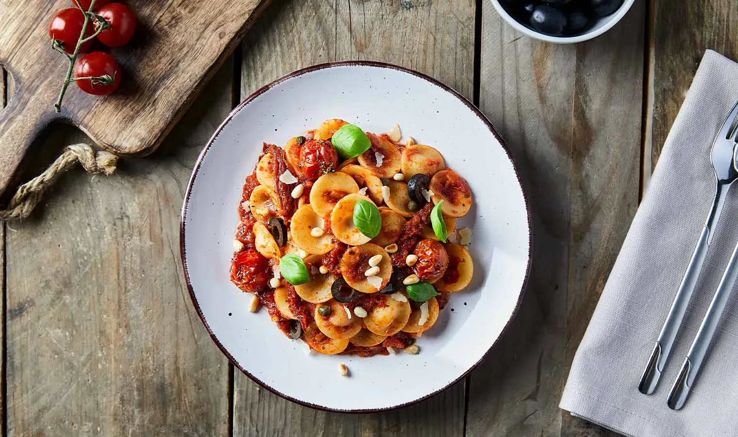 Pasta alla Puttanesca mit Orecchiette, Tomaten, Oliven und Basilikum auf einem Holztisch