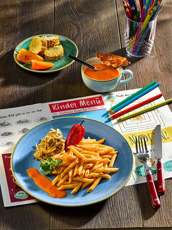 Kinderteller Pulled Chicken mit Dinkel-Penne und Paprika-Rahm, dazu Gemüse, Suppe und Buntstifte auf Holztisch
