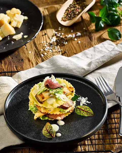 Rehlasagne mit Pilzen und Wirsing, kreativ mit Salat auf einem schwarzen Teller angerichtet, Hochformat