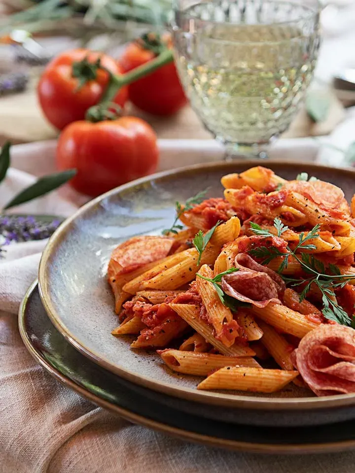 Salami-Penne in Tomatensauce, mit Kräutern garniert, auf einem Teller, im Hintergrund Tomaten