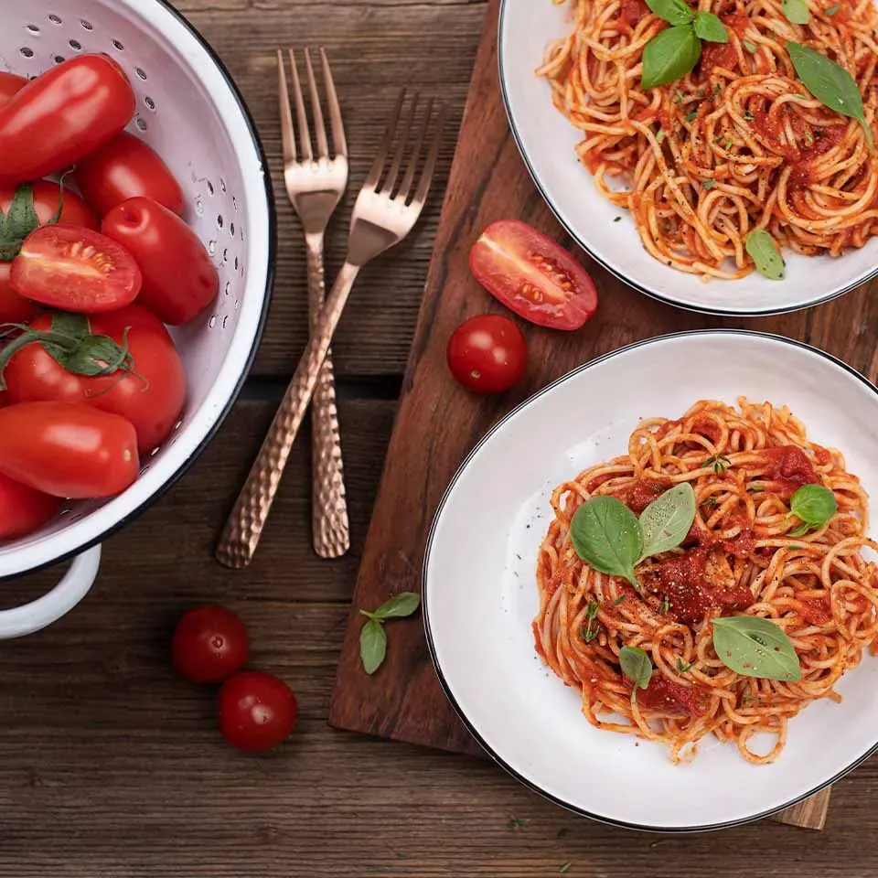 Selbstgemachte Tomatensauce zu Spaghetti auf zwei Tellern, dazu frische Tomaten in einem Sieb auf Holztisch