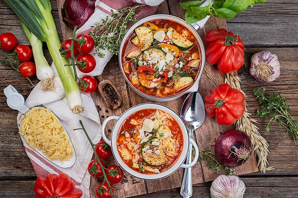 Sommer-Minestrone mit Nudelreis: zwei Schalen Gemüsesuppe mit frischem Gemüse und Kräutern auf Holztisch