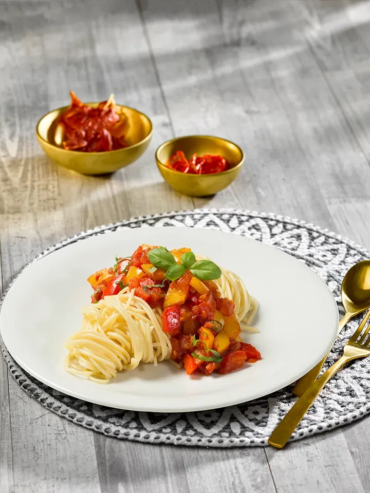 Spaghetti lunghi mit Tomaten-Paprika-Basilikum-Sauce auf grauem Tisch mit goldfarbenem Besteck
