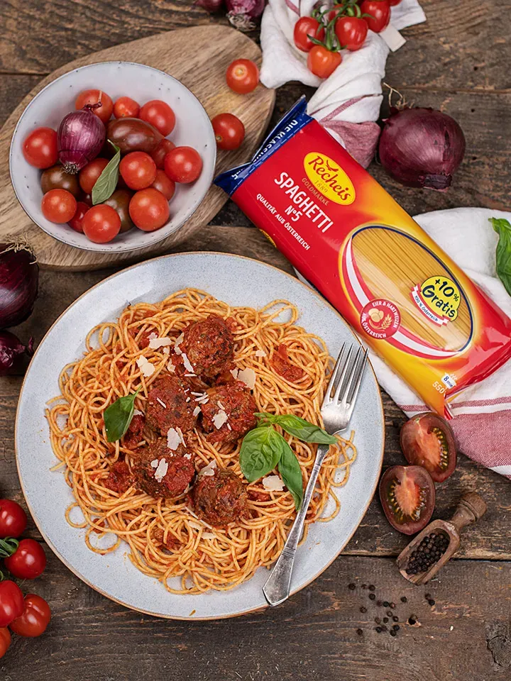 Spaghetti mit Fleischbällchen auf einem Teller, daneben Tomaten, Zwiebeln und eine Spaghettipackung