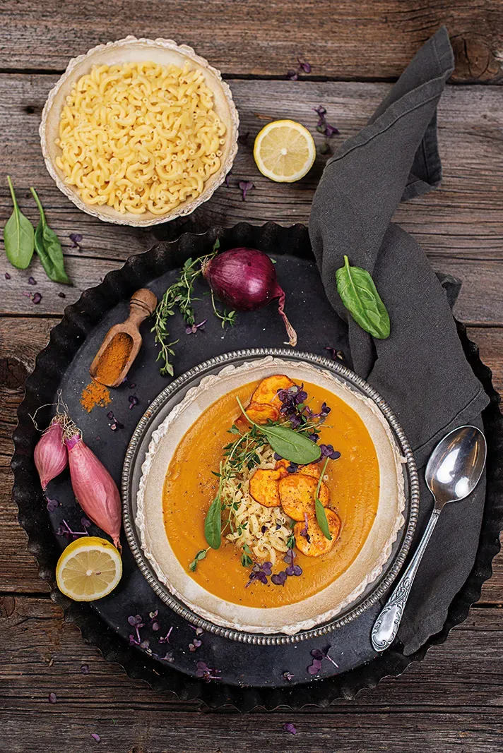 Süßkartoffel-Kurkuma-Suppe mit Pasta und Kräutern auf einem Teller auf Holz, daneben Zwiebeln und Zitrone