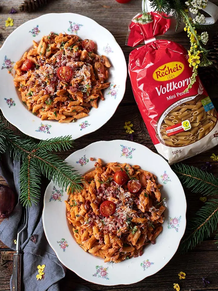 Zwei Teller Vollkorn-Mini-Penne mit Tomaten-Sahne-Speck-Sauce neben einer Packung Vollkorn-Penne