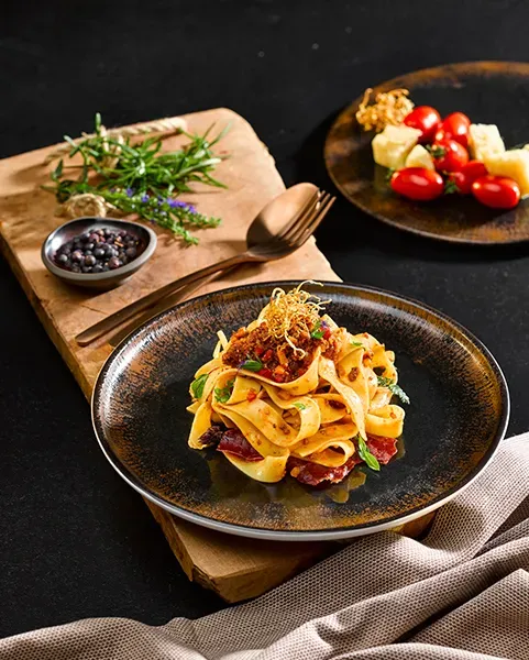 Wildbolognese mit Pappardelle auf einem Teller auf Holzbrett vor schwarzem Hintergrund, mit Kräutern und Gewürzen, im Hochformat