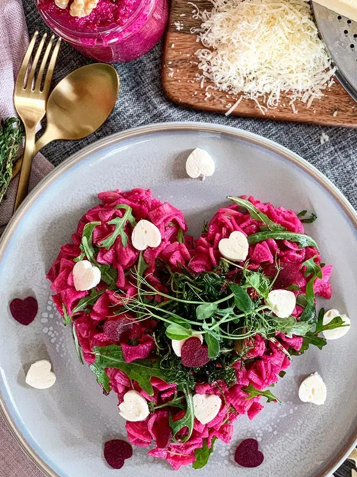 Herzförmige rote Pasta mit Rote-Rüben-Pesto, grünen Kräutern und weißen Herzen auf einem Teller, Valentinstagsrezept