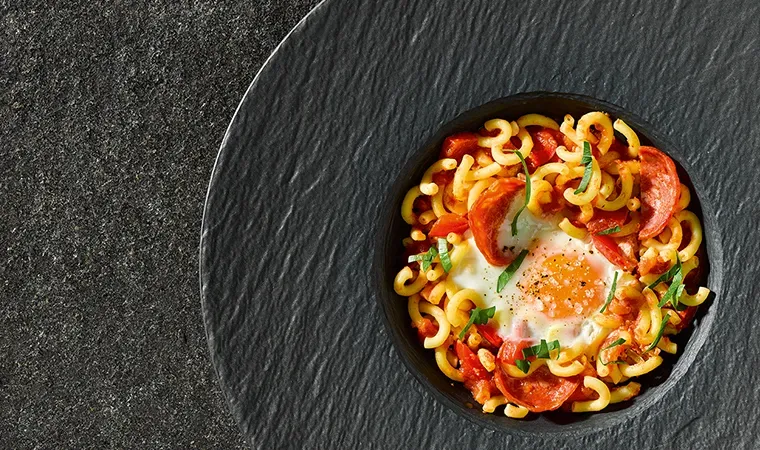 Shakshuka auf schwarzem Teller mit Nudeln, Spiegelei, Tomaten und Kräutern vor dunklem Untergrund