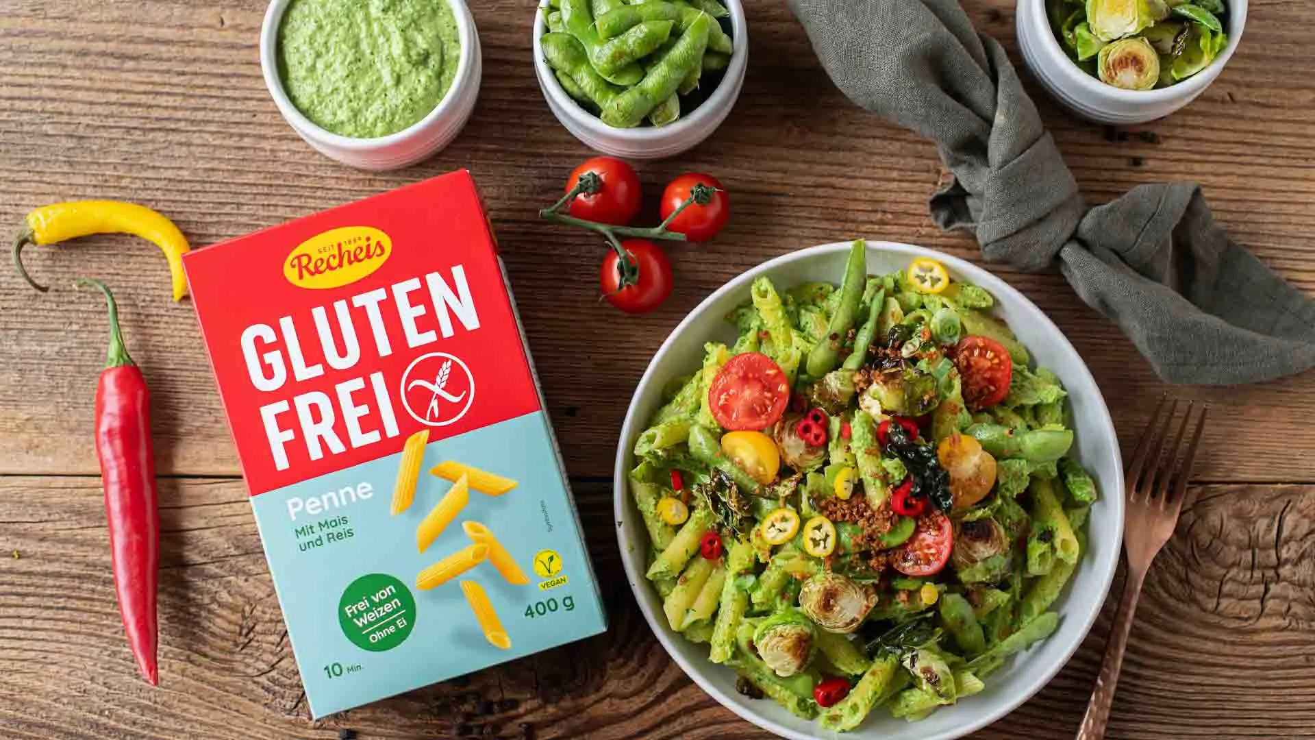 Packung Recheis Glutenfrei Penne neben einer Schüssel Penne mit grünem Pesto, Kirschtomaten und Rosenkohl