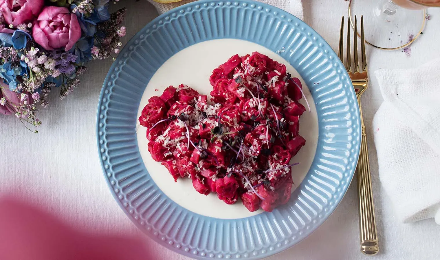 Rote Mac and Cheese in Herzform auf einem blauen Teller mit goldener Gabel, daneben ein Blumenstrauß