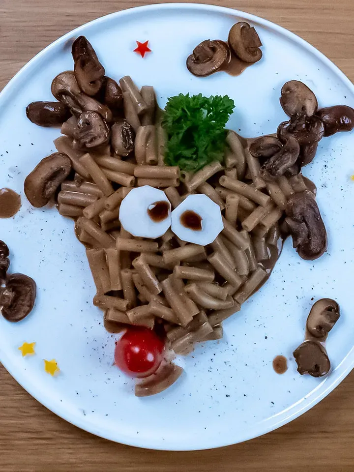 Nahaufnahme: Rudolph, das Nudeltier, aus Nudeln und Pilzen als Rentiergesicht mit Tomatennase und Petersilienhaar