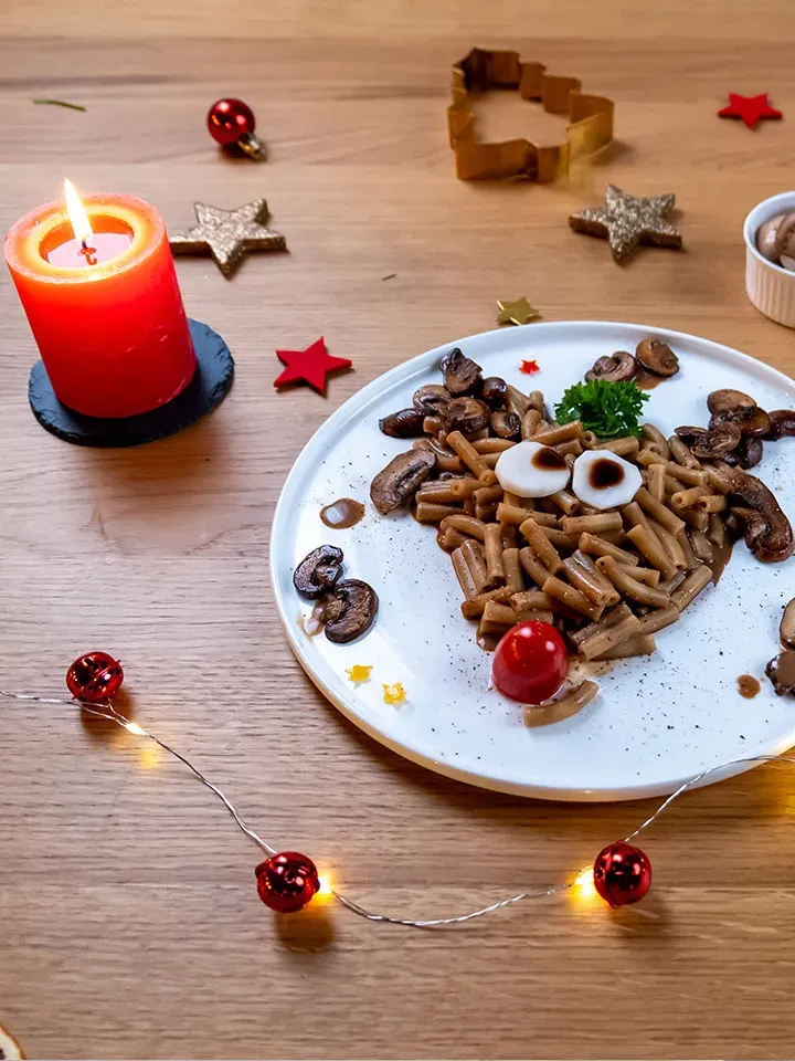 Rudolph, das Nudeltier: Pasta mit Pilzen als Rentier auf einem Teller neben brennender roter Kerze und Weihnachtsdeko