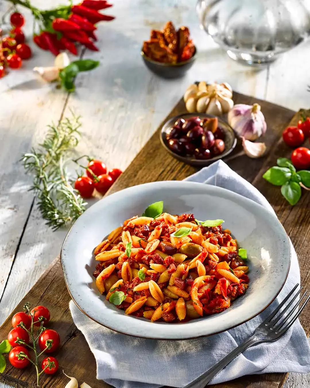 Gnocchi sardi mit Salsiccia und Peperonata in Tomatensauce, umgeben von frischen Zutaten auf einem Holztisch