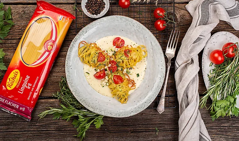 Salzburger Nudelnockerl mit Käsesauce und Tomaten auf einem Teller, daneben Nudelverpackung und Kräuter auf Holztisch