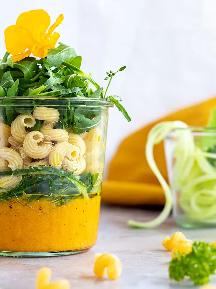 Schichtsalat im Glas zum Mitnehmen mit Nudeln, grünem Salat und gelber Sauce, mit Blume garniert