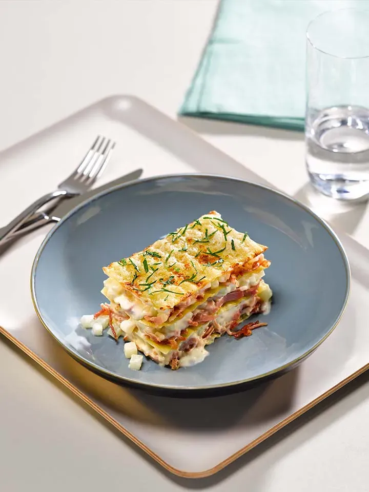 Nahaufnahme Schinken-Kohlrabi-Lasagne: ein Stück auf blauem Teller, Besteck und Glas Wasser auf Tablett