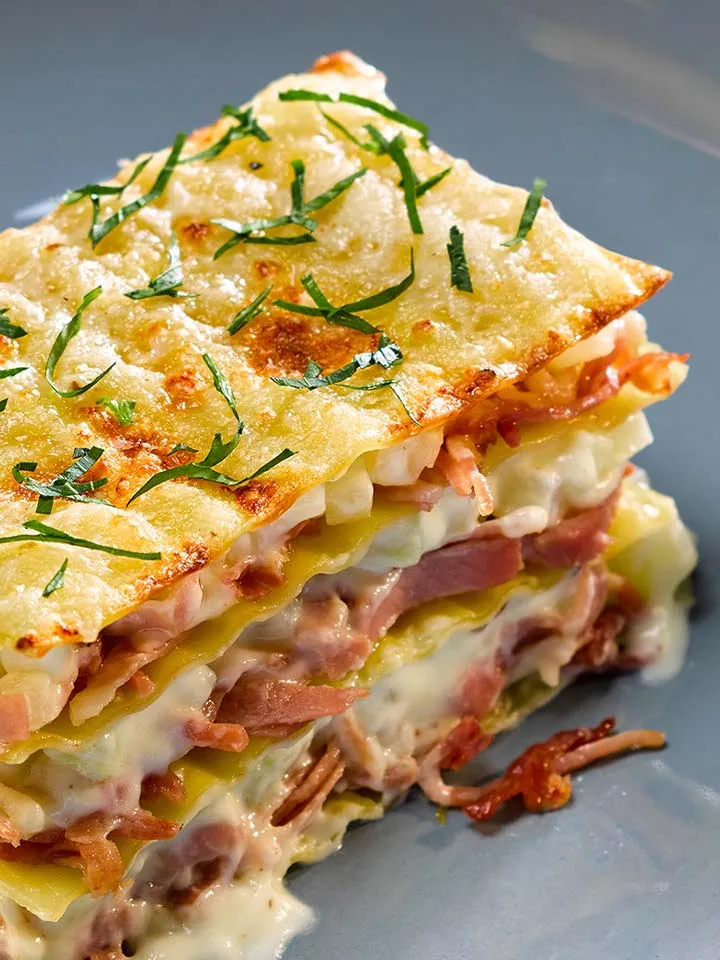 Nahaufnahme Schinken-Kohlrabi-Lasagne: ein Stück auf blauem Teller mit Besteck und Glas Wasser