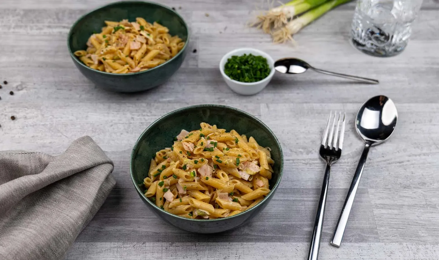 Schinken-Lauch-Nudeln mit Penne und Kräutern in zwei Schalen auf grauem Tisch mit Besteck