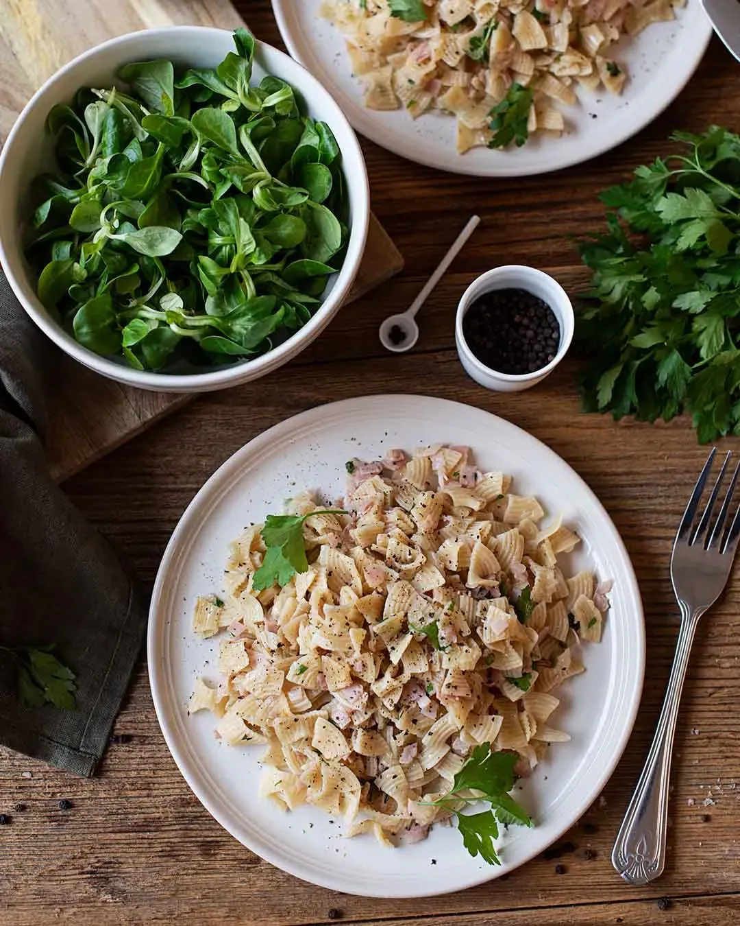 Schinkenfleckerl auf Teller, daneben Schüssel mit grünem Salat und Gewürzen auf Holztisch