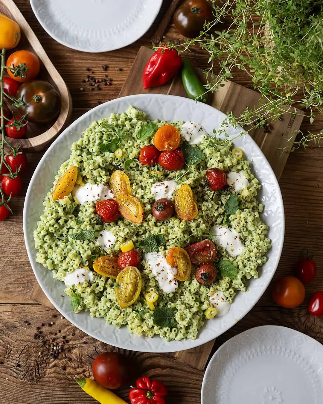 Aufsicht auf eine Schale Joghurt-Nudeln mit Ofentomaten auf einem Holztisch mit Tomaten, Paprika und Kräutern