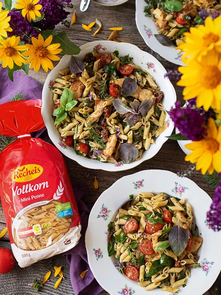 Sommernudelsalat mit Vollkorn-Mini-Penne und Hühnerfilets auf zwei Tellern, daneben bunte Blumen auf Holztisch