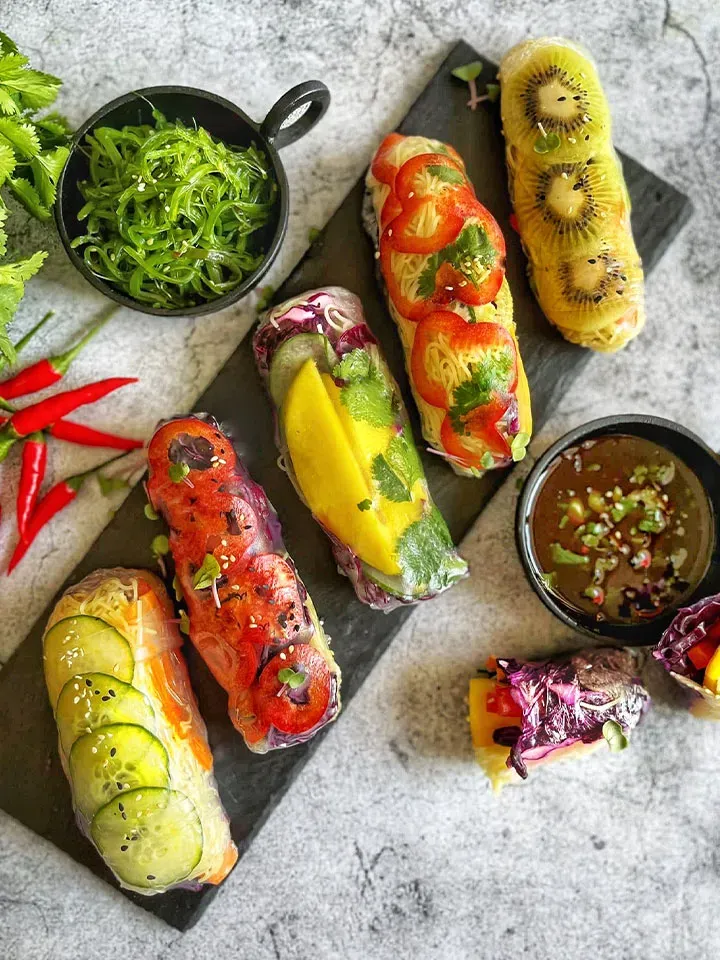 Bunte vegane Sommerrollen mit Nudeln, Gemüse und Früchten auf einem schwarzen Brett mit Dip
