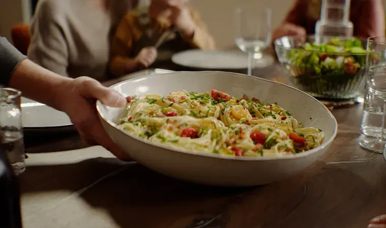 Person reicht eine Schüssel Spaghetti Carbonara mit Tomaten am gedeckten Tisch