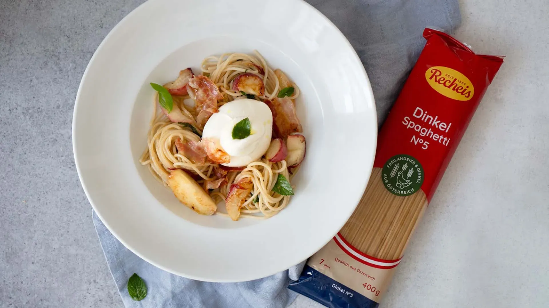 Spaghetti mit gebratenem Pfirsich und Burrata, dazu Ei und Speck, neben einer Packung Dinkelspaghetti