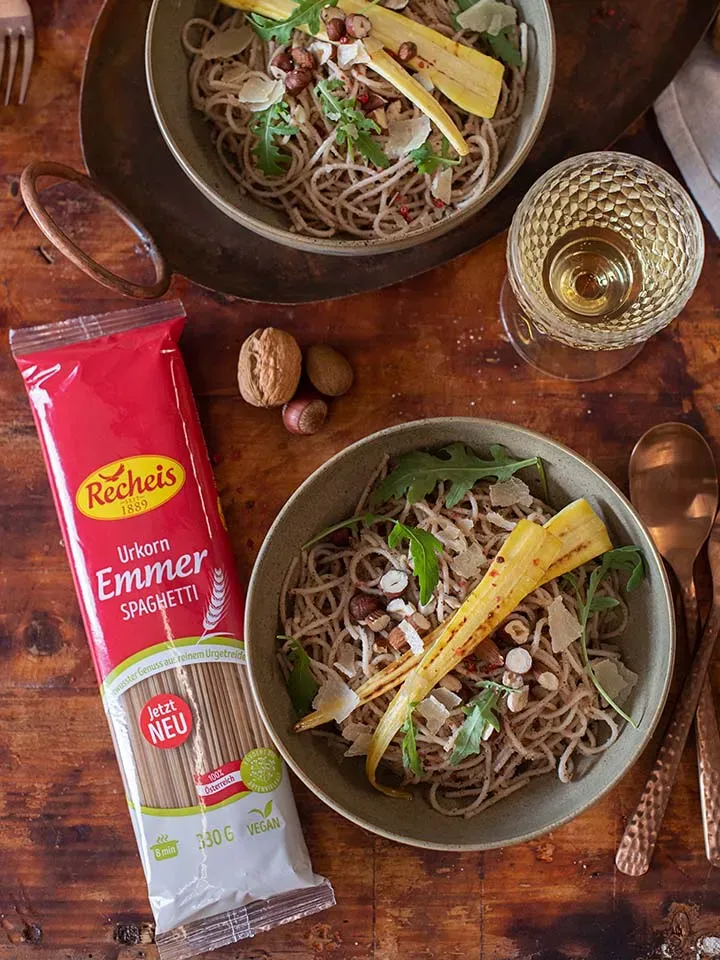 Zwei Schalen Emmer-Spaghetti mit Nusspesto, Rucola und Petersilienwurzel auf Holztisch, dazu Pastapackung und Weinglas